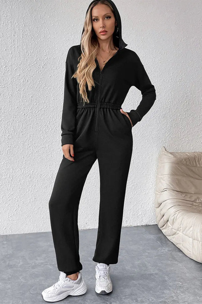 Woman wearing a black jumpsuit standing against a white wall with a beige chair.