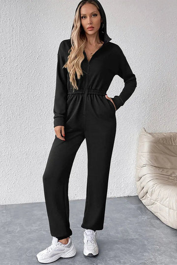Woman wearing a black jumpsuit standing against a white wall with a beige chair.